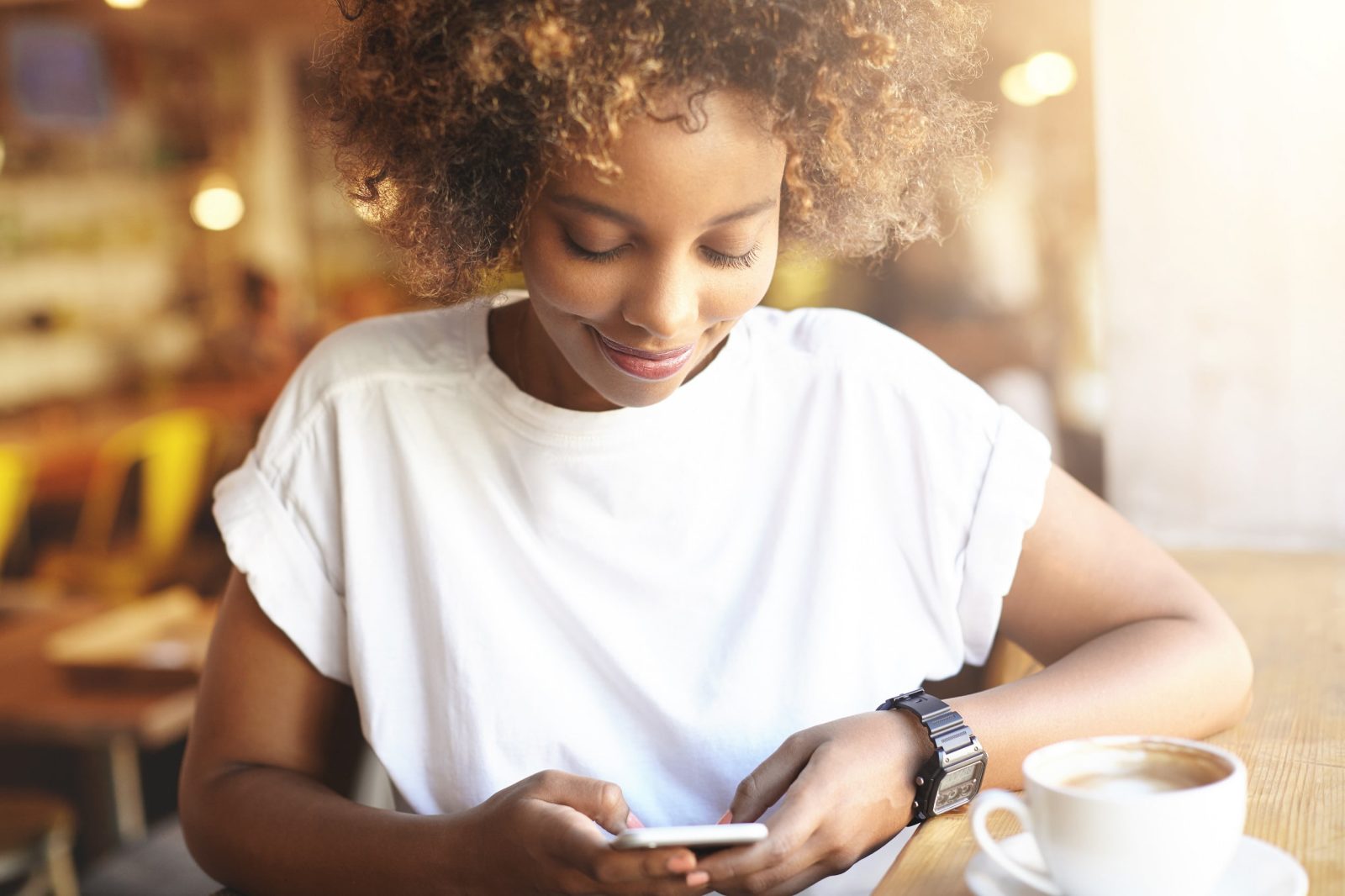Portrait of fashionable African brunette female with Afro haircut using high-speed Internet connection at cafe, checking her news feed with joyful smile, sitting at cafe with cup of coffee on table